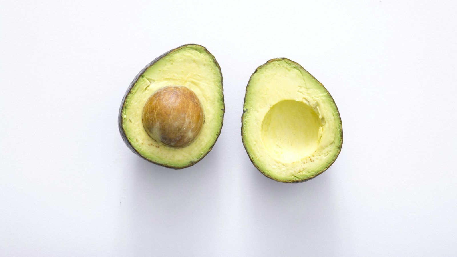 An avocado cut in half on a white surface, with the pit in one half.