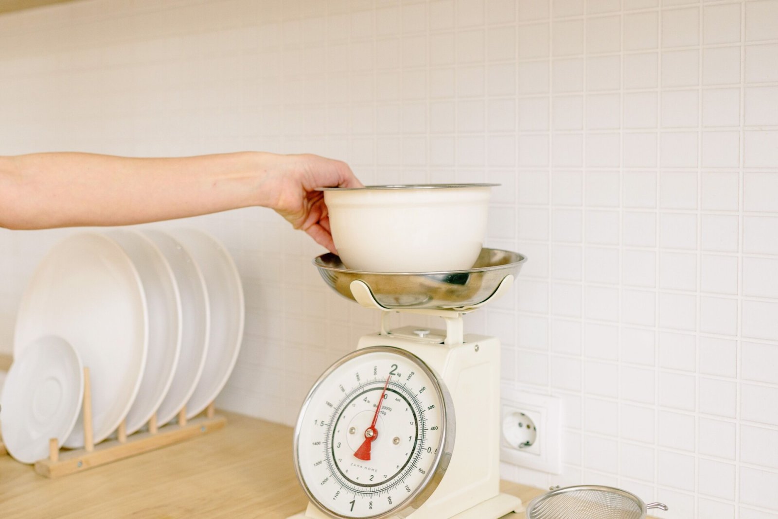 A hand places a white bowl on a kitchen scale for food preparation.