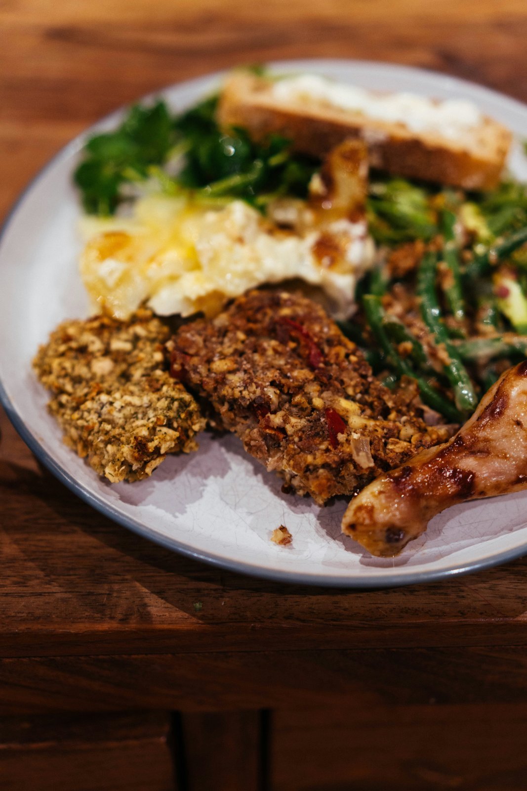 A dinner plate with a chicken drumstick, mashed potatoes, green vegetables and slices of a baked loaf on a wooden table.