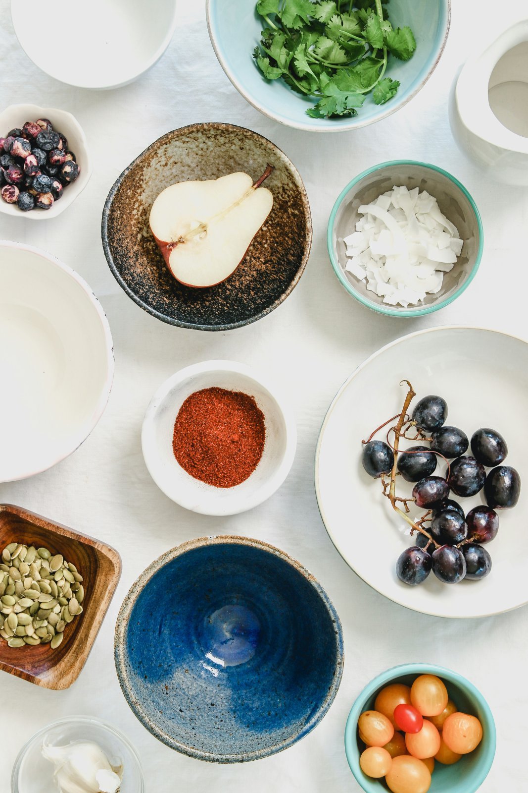 Overhead view of bowls holding a halved pear, grapes, pumpkin seeds, coconut flakes, herbs and cherry tomatoes on a white tablecloth.